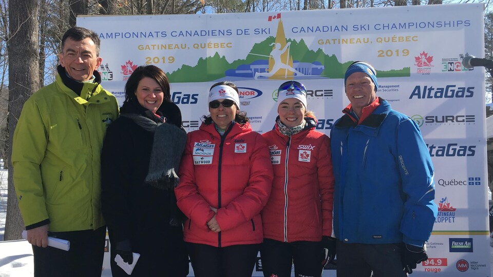 Cinq personnes posent pour la caméra devant une affiche qui annonce la tenue des championnats canadiens de ski de fond à Cantley.