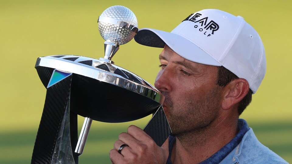 Un homme embrasse un trophée sur lequel se trouve une balle de golf argentée.