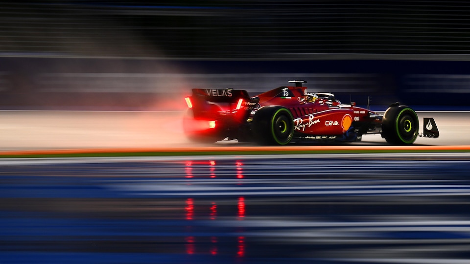 Une monoplace de F1, vue de profil, roule sur un circuit mouillé. On voit le reflet de ses lumières rouges dans les flaques d'eau.