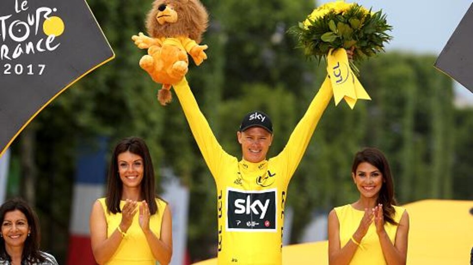 Un coureur en jaune tout sourire, accompagné de deux hôtesses sur un podium.