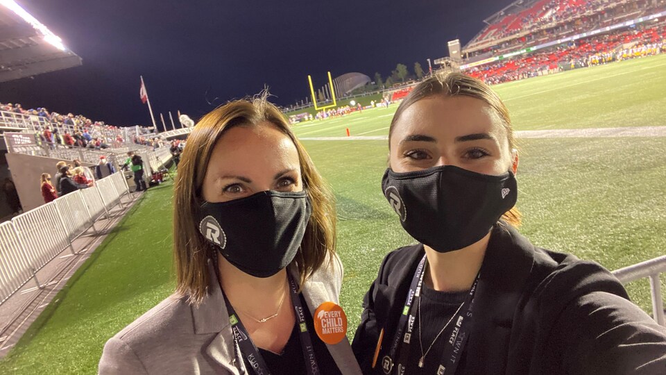 Two women take a selfie in front of a football field. 