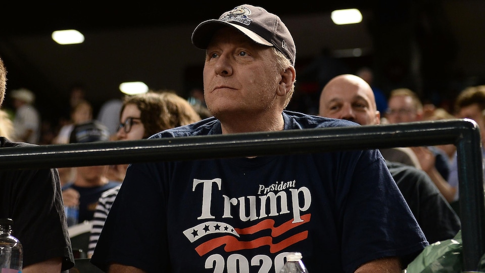 Un homme avec un chandail bleu soutenant Donald Trump dans les gradins.