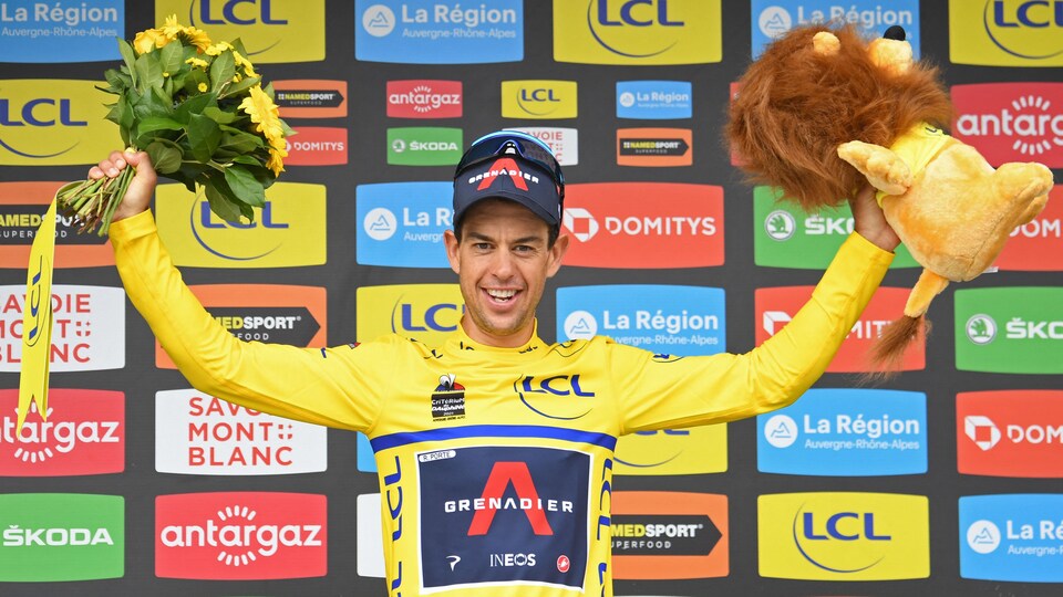 Il sourit et lève les bras, un lion en peluche dans la main gauche et un bouquet de fleurs dans la main droite.