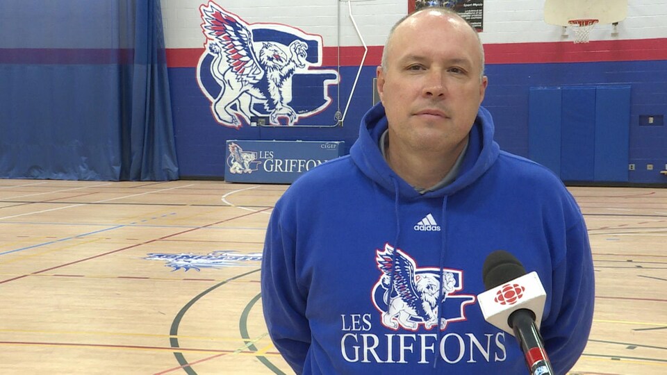 Un homme est en entrevue dans un gymnase avec un gros logo de l'équipe sportive locale en arrière-plan.