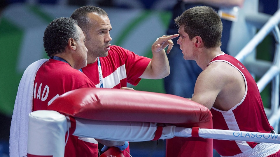 Un entraîneur de boxe, vêtu de rouge, prodigue des conseils en pointant son index sur le front de son athlète dans un ring. 