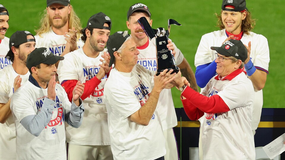 Un homme coiffé d'une casquette tient un trophée alors que des joueurs de baseball l'applaudissent.