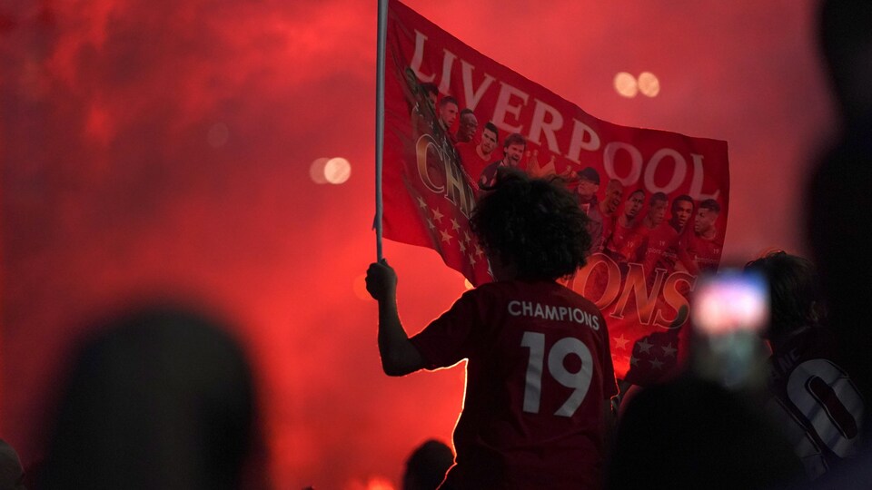 Un homme assis sur les épaules d'un autre porte un t-shirt avec le no 19 et le mot "champions". Il tient aussi un drapeau de Liverpool dans sa main.