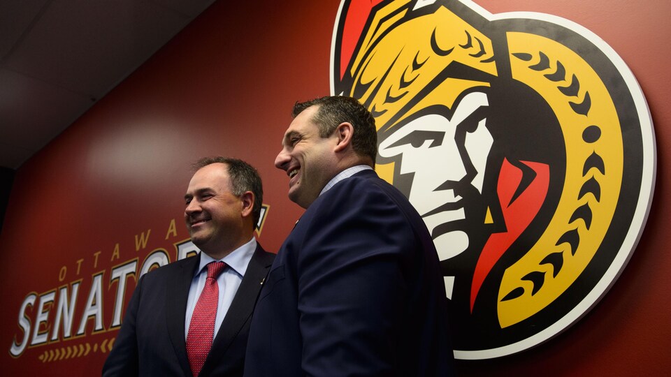Pierre Dorion pose avec D.J. Smith devant un gros logo des Sénateurs.