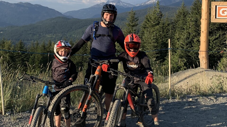 Un homme se tient entre ses deux enfants qui enfourchent un vélo sur un sentier en montagne.