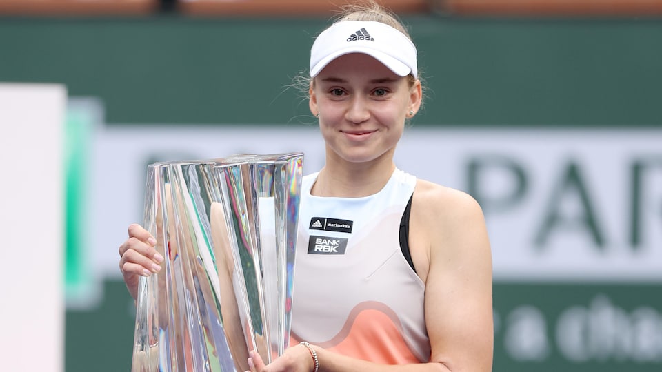 Une joueuse de tennis tient un trophée en verre et sourit.