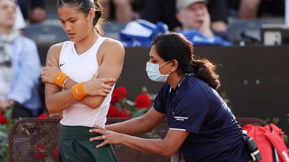 Female tennis player with arms crossed, masked physiotherapist with lower back mass on court.