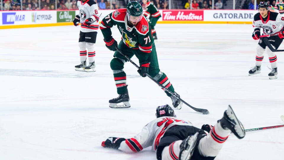 Joueur de hockey, maniant la rondelle, devant un adversaire qui est couché sur la glace.
