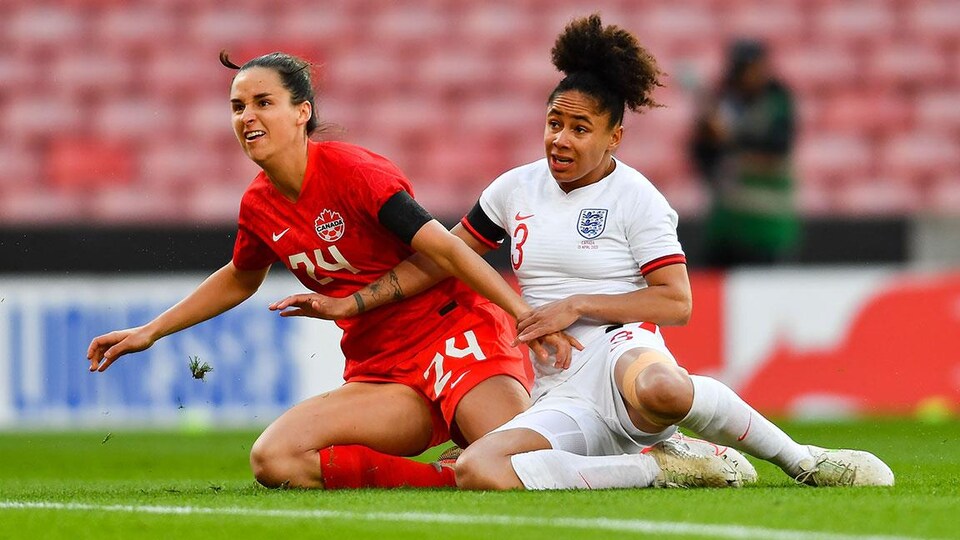 L'attaquante québécoise, à genoux au sol avec une défenseuse de l'Angleterre, regarde l'action se poursuivre.