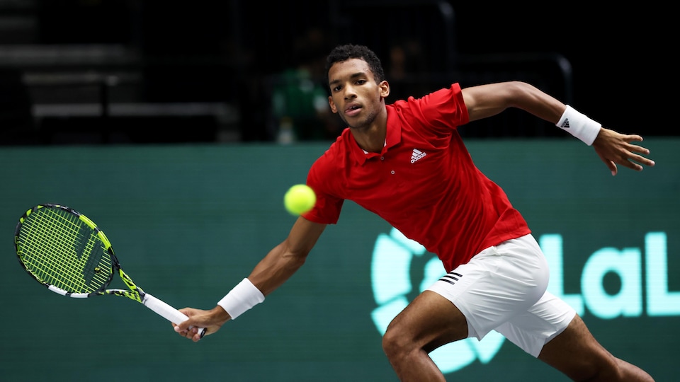 A tennis player, wearing a red sweater and white shorts, stretches to hit a forehand ball with his yellow racquet. 