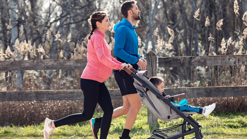 Une femme et un homme font de la course à pied. La femme est enceinte et pousse un enfant dans une poussette. 