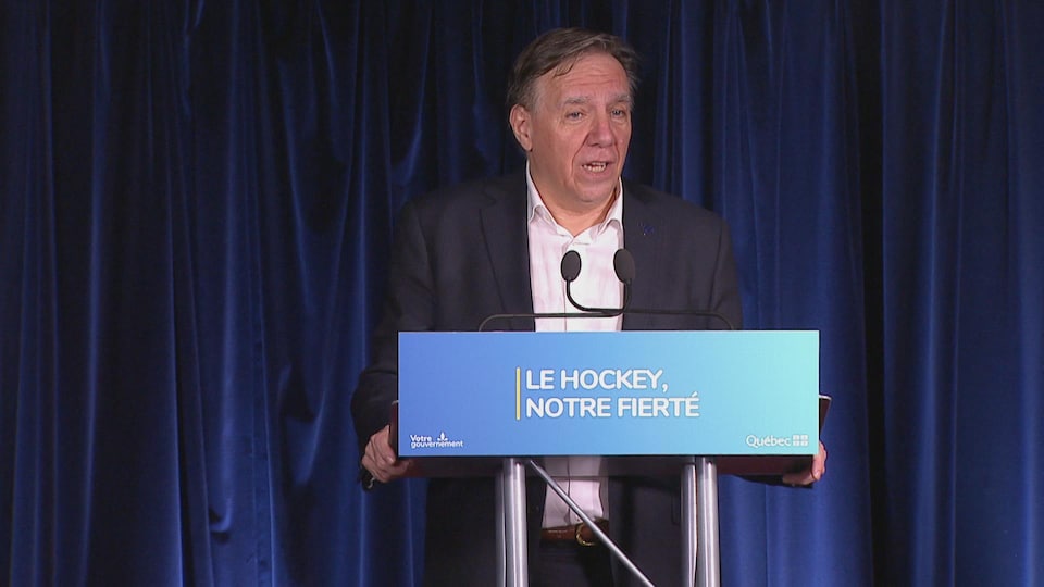 Le premier ministre du Québec, en veston, parle pendant une conférence de presse, devant un podium avec les mots «Le hockey, notre fierté».