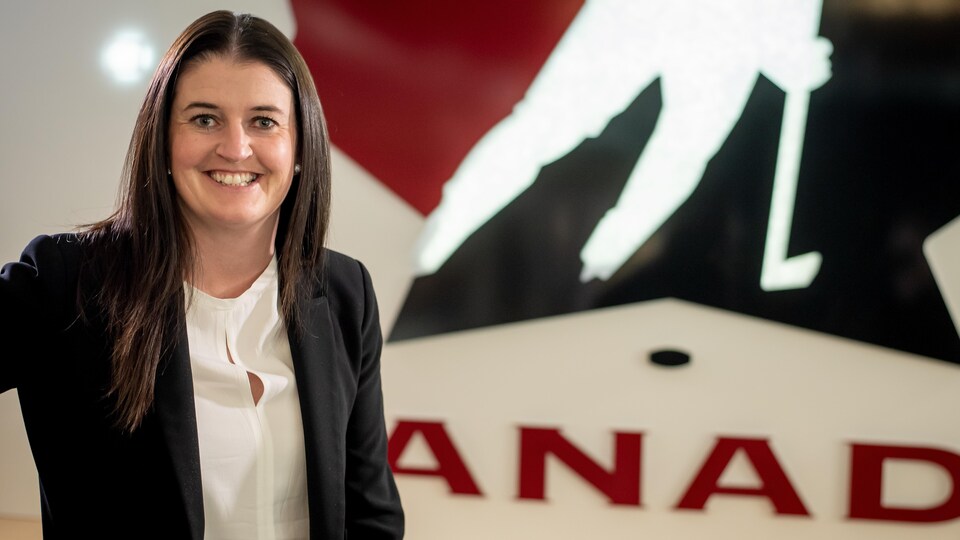 Une femme vêtue d'un veston noire et d'une chemise blanche prend la pose devant le logo de Hockey Canada.
