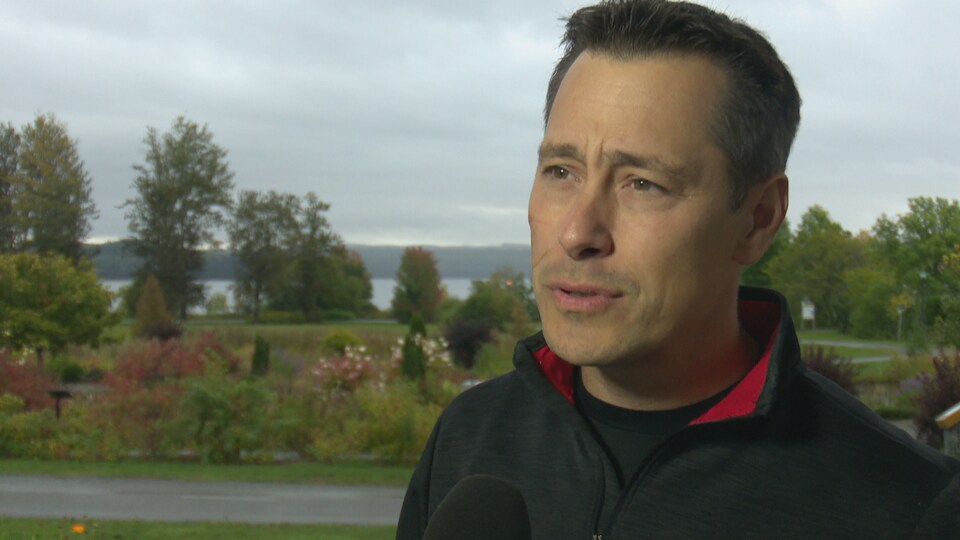 Plan buste de Guy Boucher en entrevue avec le parterre de sa résidence en arrière-plan
