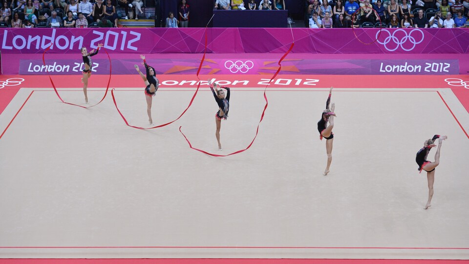 Cinq gymnastes se tiennent une jambe, trois d'entre elles ont lancé un ruban rouge en l'air.