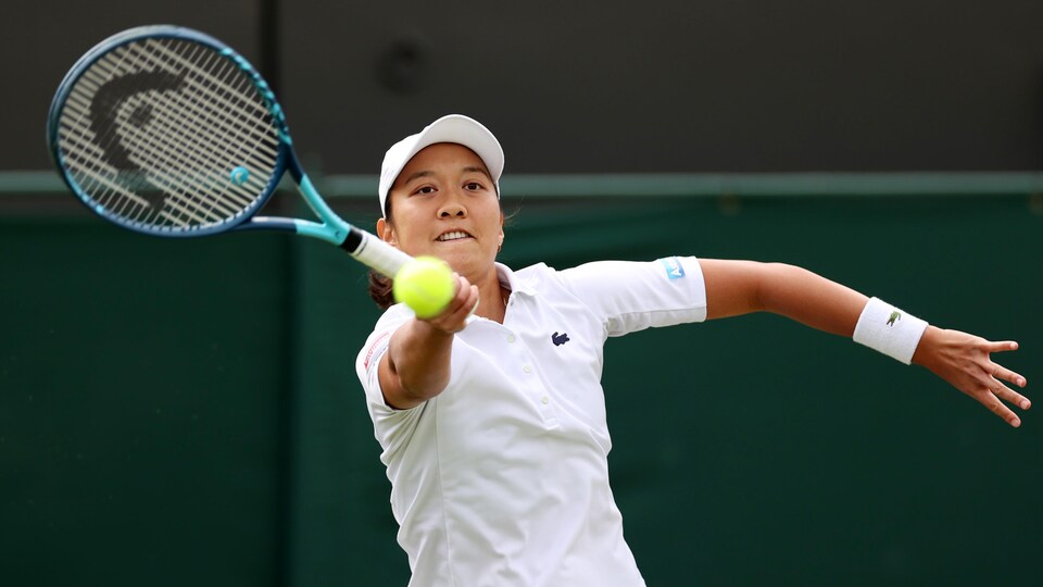 La joueuse de tennis, en blanc, une casquette sur la tête, se concentre sur la balle devant elle. 