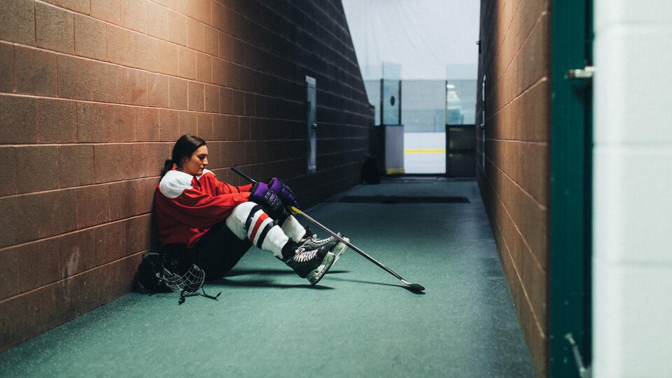 Une joueuse de hockey est assise par terre dans un corridor menant à la glace. Elle semble triste.