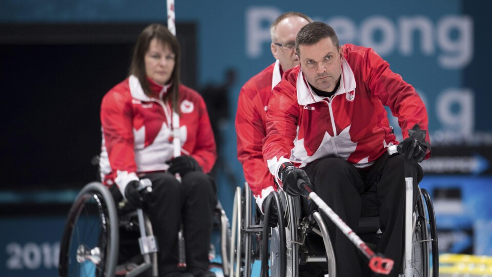 Des joueurs de curling sont concentrés pendant un match. Le joueur en avant-plan tient un balai.