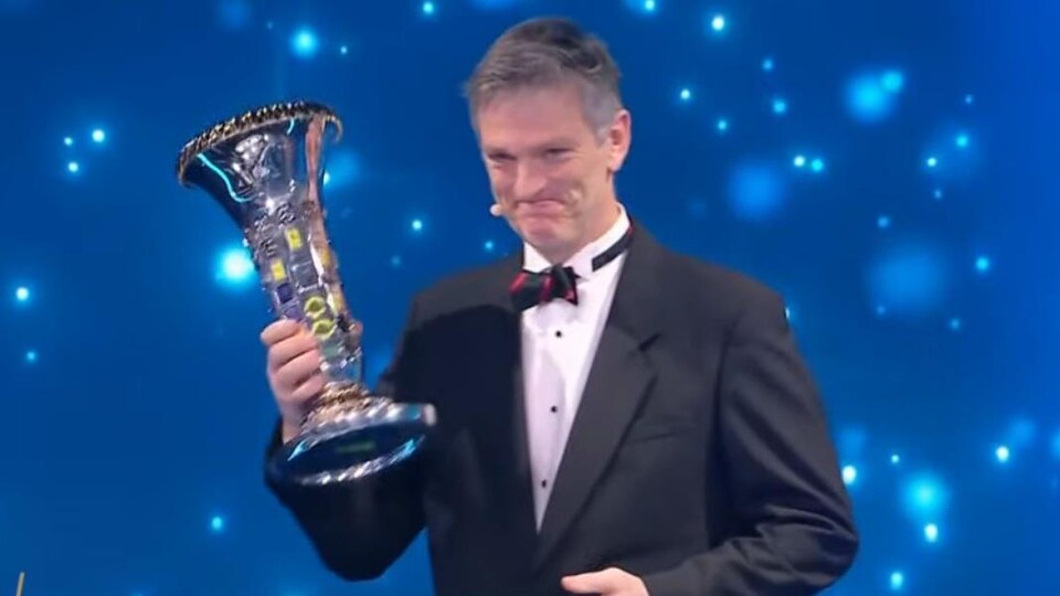 Un homme en habit de soirée sourit avec un trophée dans la main droite.