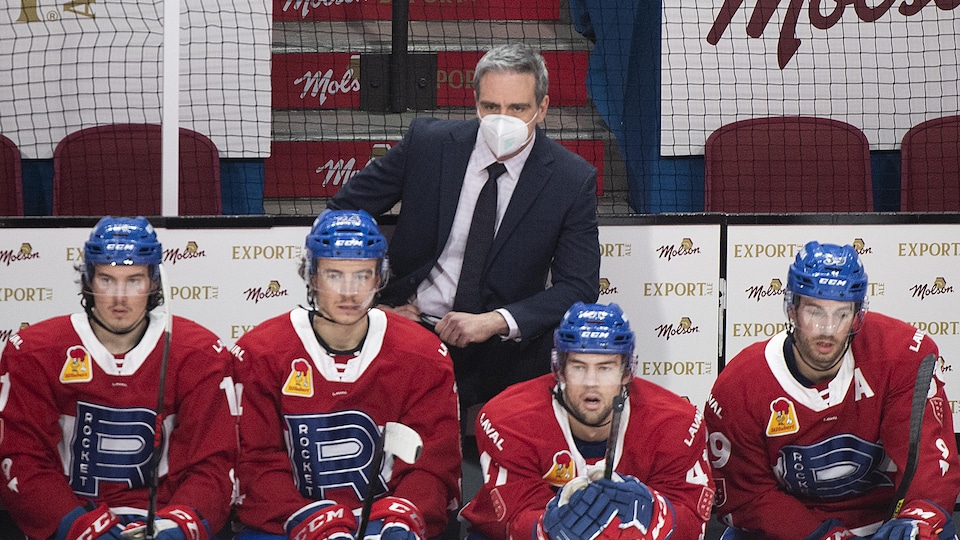 L'entraîneur-chef, debout derrière le banc des joueurs, regarde l'action sur la glace.