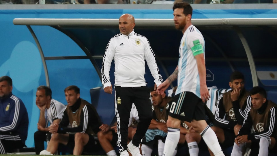 Un joueur de soccer marche sur le terrain, on voit son entraîneur sur la touche et ses coéquipiers dans l'abri.