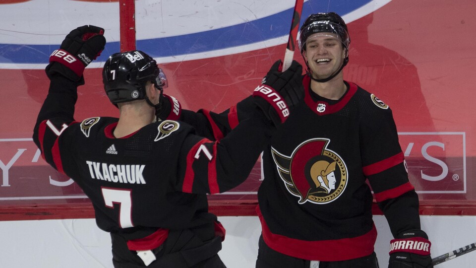 Le joueur de centre sourit, accoté contre la bande, et accueille son coéquipier Brady Tkachuk pour célébrer son but en prolongation.