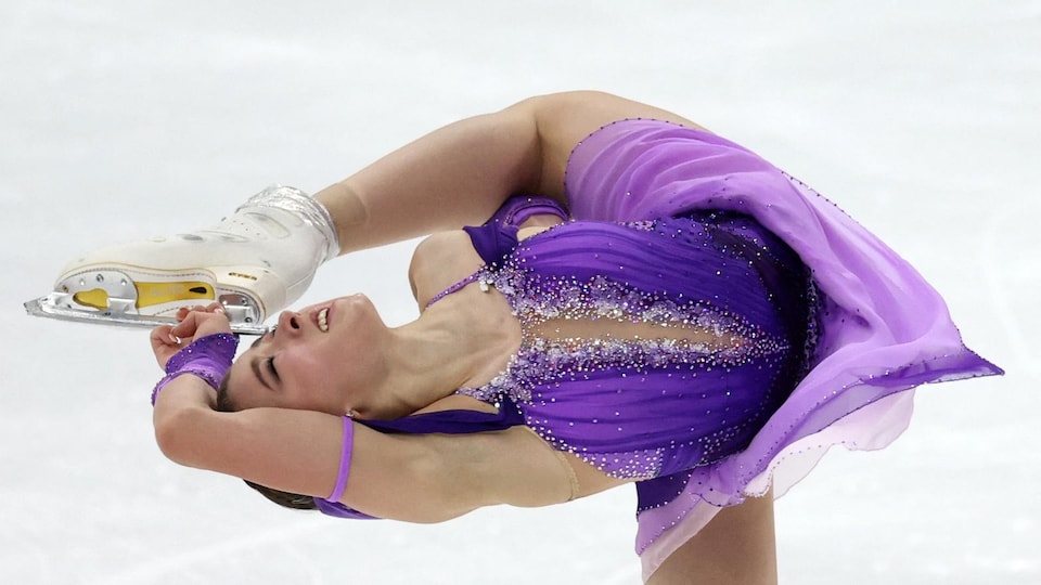 Une patineuse réalise une figure, le dos perpendiculaire à la glace en tenant don patin gauche derrière sa tête.