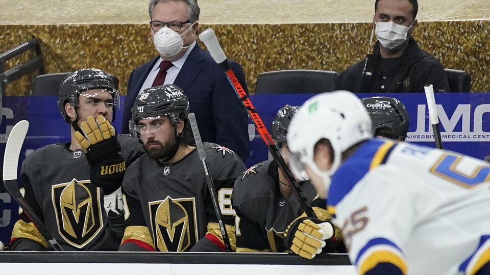 Masqué, il dirige son équipe derrière le banc des joueurs.