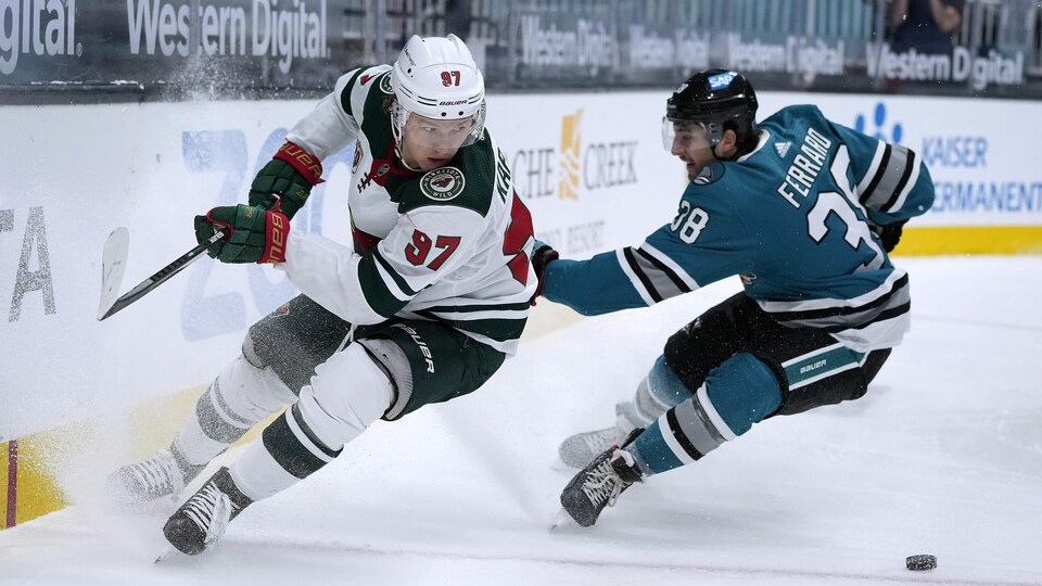 L'attaquant du Wild, vêtu de l'uniforme blanc, freine brusquement et cherche la rondelle derrière lui, alors que le défenseur Mario Ferraro, des Sharks, lui met une main dans le dos, lors d'un match à San José.