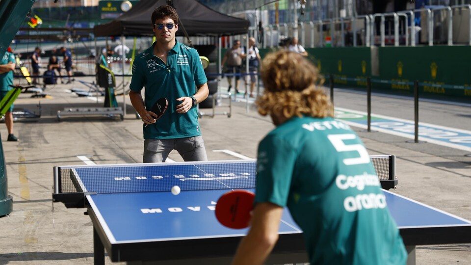 Deux pilotes de F1 jouent un match de tennis de table dans les puits d'un circuit.