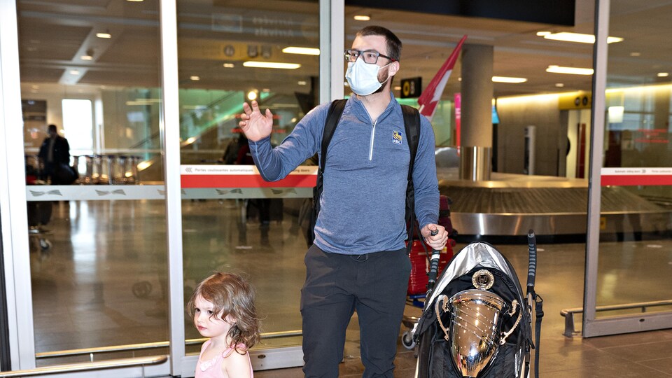 Un homme et une enfant se tiennent à côté d'une poussette où se trouve un trophée. Ils sont à l'aéroport.
