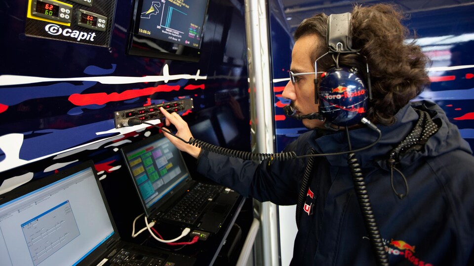 Un homme casqué, de profil, appuie sur un bouton sur un mur d'écrans dans un garage d'une équipe de F1.