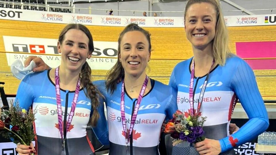 Trois cyclistes canadiennes en combinaison sourient pour la photo avec leurs médailles devant une piste.