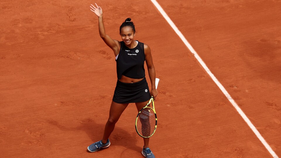 A tennis player dressed in black waves in front of the crowd after her victory on the red clay court of the Roland-Garros tournament. 