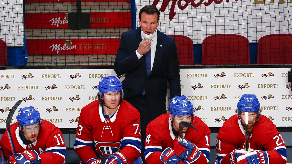 Vêtu d'un complet, debout derrière le banc du Canadien, l'entraîneur retient son masque sous son menton pour donner des instructions aux joueurs sur la glace.
