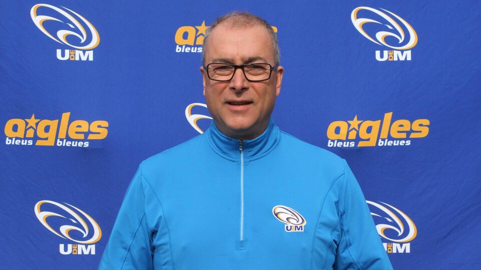 Homme avec lunettes et cheveux courts devant un drap bleu avec des logos des Aigles Bleus.
