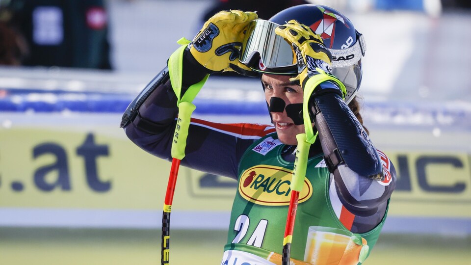 Une skieuse retire ses lunettes dans l'aire d'arrivée d'une épreuve, ses bâtons accrochés aux poignets.