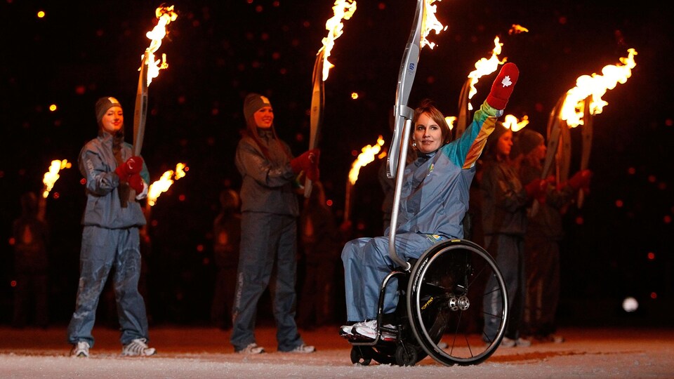 Een vrouw in een rolstoel zwaait naar de menigte.  Aan zijn stoel is een olympische fakkel bevestigd.