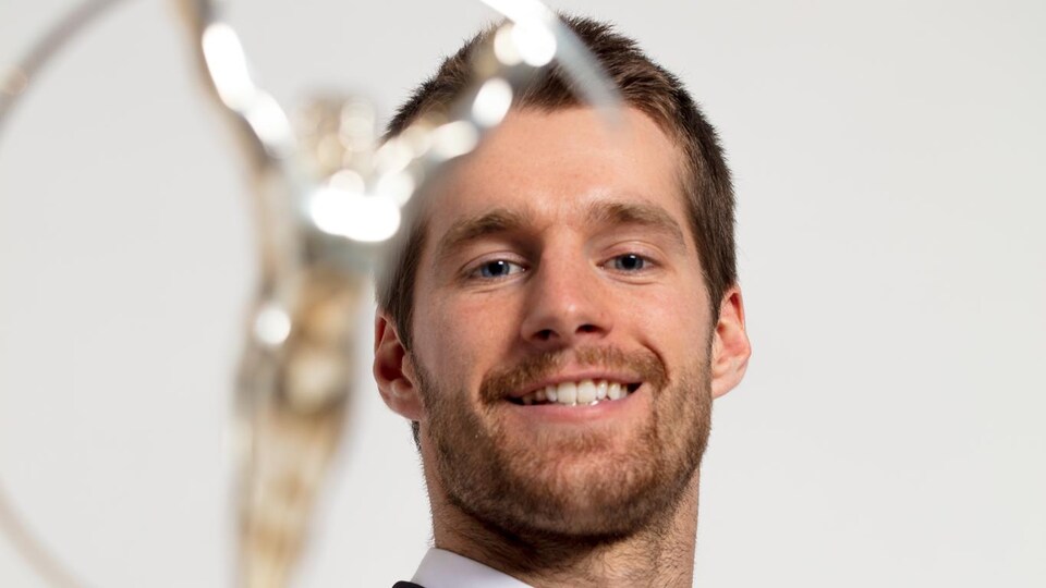 Il sourit en tenant son trophée à bout de bras, devant lui.