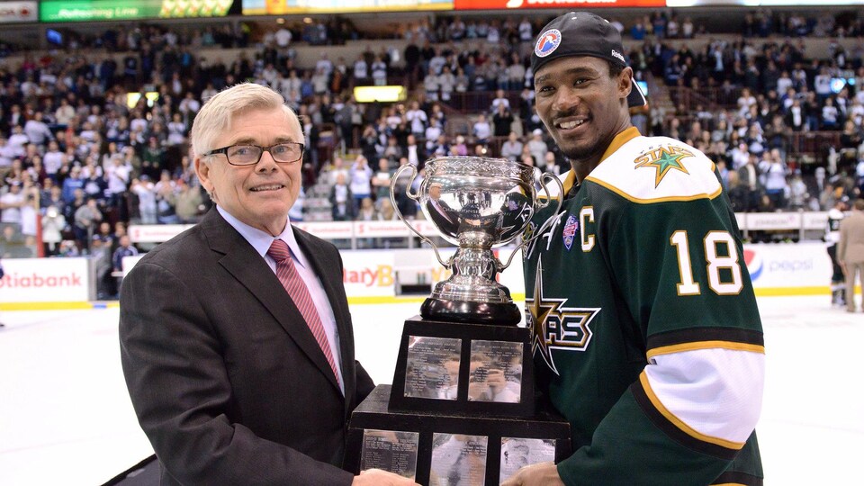 Un joueur de hockey vêtu d'un uniforme vert reçoit un trophée des mains d'un dignitaire au centre de la glace. 