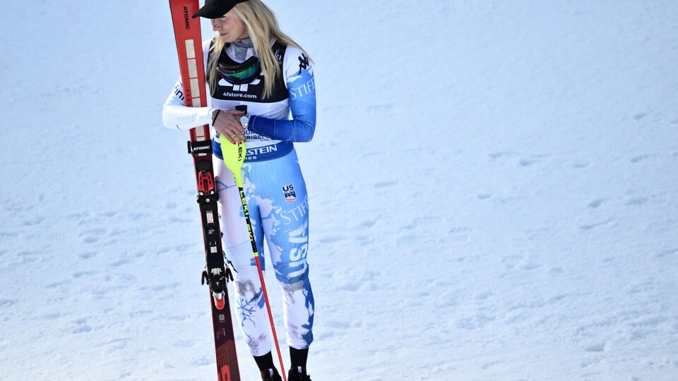 Une skieuse debout dans la neige tient ses skis et ses bâtons, regarde sur sa droite et sourit.  