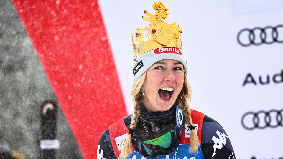 Une skieuse souriante fait gicler le champagne, avec une couronne sur la tête et une médaille d'or autour du cou. 