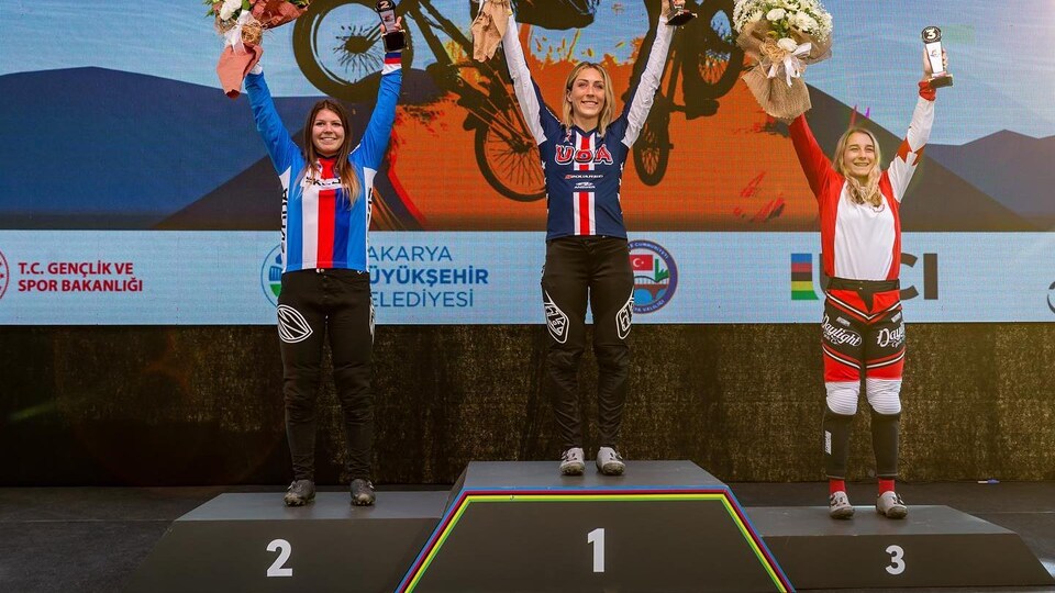 Molly Simpson est sur la troisième marche du podium, les bras dans les airs avec un bouquet dans la main droite et un trophée dans la main gauche.