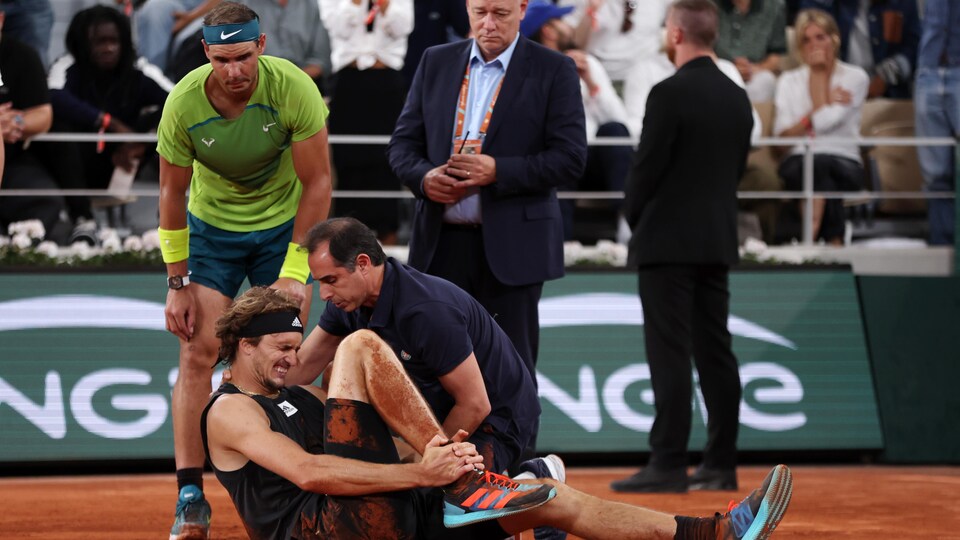 Un joueur de tennis se tord de douleur au sol. Son adversaire, à ses côtés, le regarde.