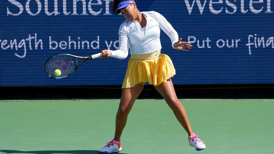 Une joueuse de tennis, vêtue d'une jupe jaune et d'un chandail blanc, frappe une balle du coup droit lors d'un match sur un terrain vert. 