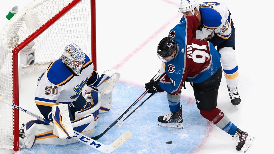 Two hockey players compete for the puck in front of a goalie who is kneeling in front of his net.  